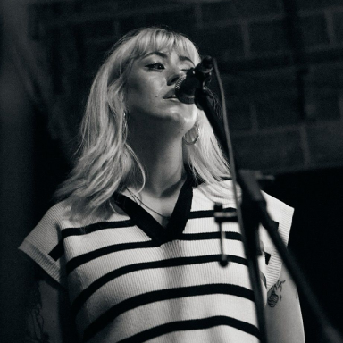 a blonde woman wears a stripy vest and sings into a mic